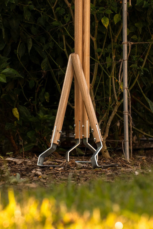 A multifunctional cultivating tool with a wooden handle and a bottom-mounted metal blade, standing upright in the ground.