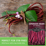 A bundle of long, thin red asparagus beans with green leaves and a small pink flower, arranged on a rustic wooden surface next to a seed packet labeled 'Asparagus Beans Red Noodle' by Survival Garden Seeds. A green banner at the bottom reads 'Perfect for stir-fries' and 'Mild flavor with crisp-tender bite.