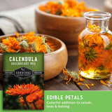 Seed packet for Calendula Greenheart Mix by Survival Garden Seeds, shown beside a glass bottle filled with orange edible petals and bowls of dried marigolds on a wooden surface.
