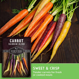 A bundle of fresh rainbow carrots in orange, yellow, red, and purple on a dark wooden surface, next to a seed packet labeled 'Carrot Rainbow Blend' by Survival Garden Seeds, with a green banner at the bottom reading 'Sweet & Crisp'.