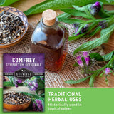 A glass bottle with a cork stopper filled with amber-brown herbal liquid sits on a burlap surface with green leaves and purple flowers around it. A bowl of dried herbs is at left, and a printed card reading 'COMFREY Symphytum Officinale' and 'Survival Garden Seeds' is overlaid.