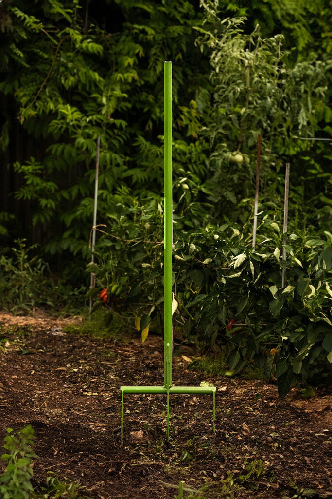 A green metal broadfork tool standing upright in a garden setting, with no visible handles or operators.