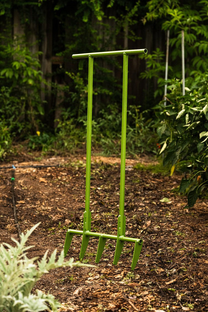 A green metal broadfork tool with spaced tines, standing upright in a garden setting.