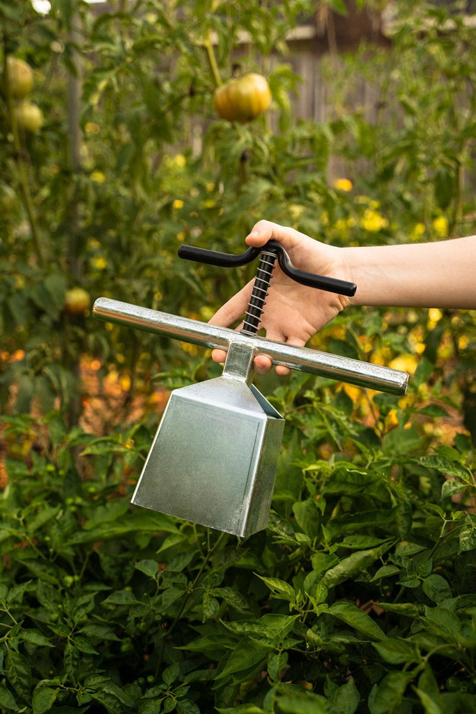 A hand holding a silver soil blocker tool in a garden with plants in the background.