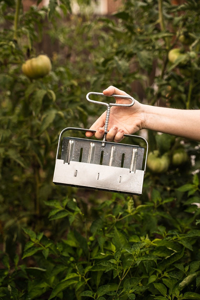 A hand holding a silver-colored soil blocker with four compartments, with plants in the background.