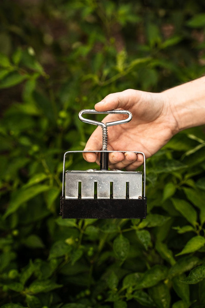 A hand holding a black soil blocker with a metal handle, against a blurred background of greenery.
