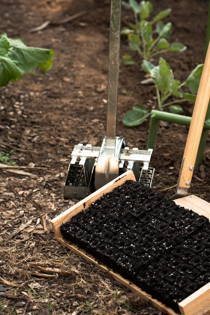 A soil blocker tool being used to shape soil in a garden setting, with a tray of soil blocks in the background.