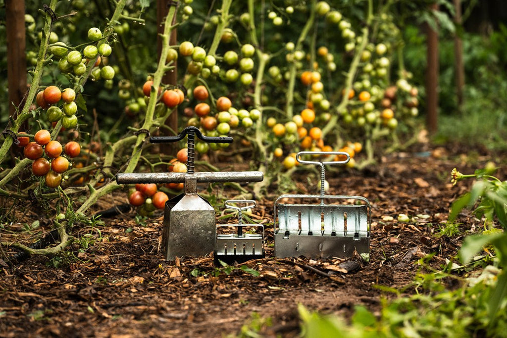 Soil blockers tools are placed in a garden setting with mature plants in the background. The tools are used for creating structured blocks in the soil, likely for supporting seedlings.