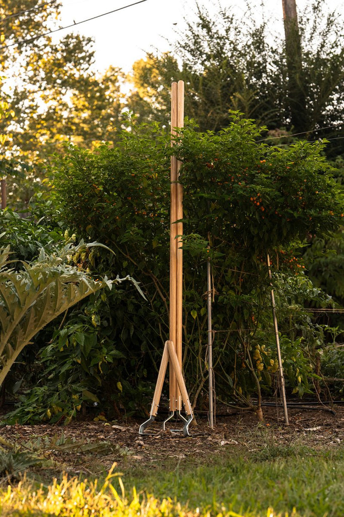 A multifunctional cultivating tool with a wooden handle and a bottom-mounted metal blade, standing upright in the ground.